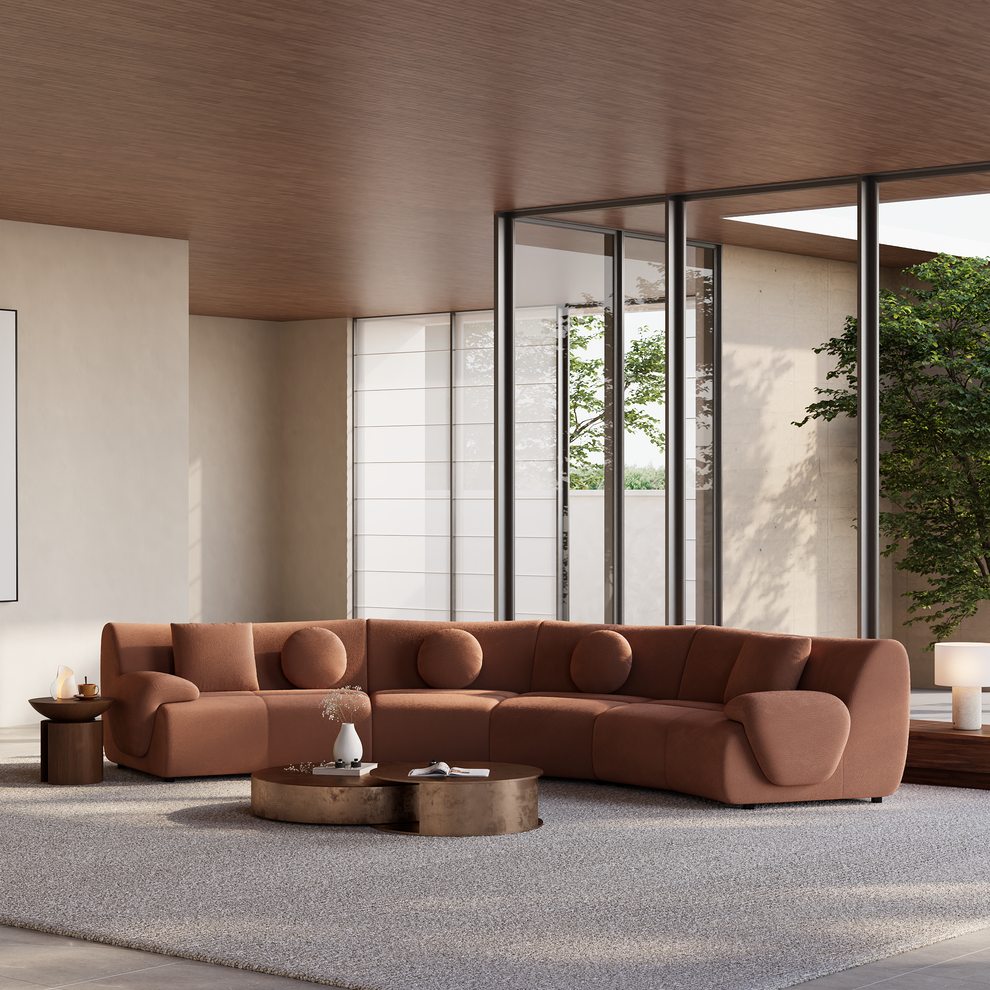 Curved rust-colored sofa with round and rectangular cushions in a bright living room with low coffee tables, a pale rug, and tall windows overlooking trees.