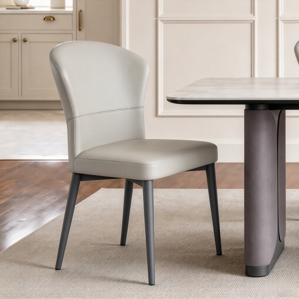 A light gray upholstered dining chair with a curved back and dark legs stands beside a table in a bright room.