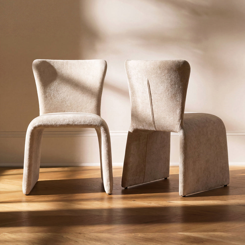 Two beige upholstered dining chairs with broad panel legs shown from the front and rear