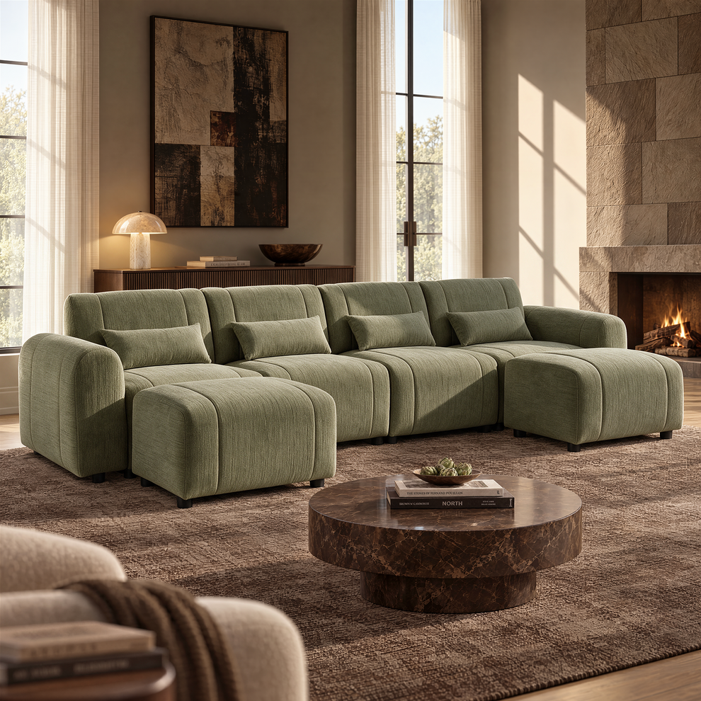 Green upholstered sofa with four matching cushions and two ottomans in a sunlit living room with a round brown coffee table, abstract wall art and a lit fireplace.
