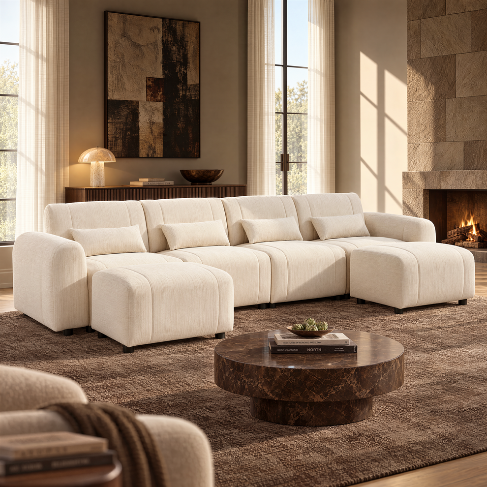 Cream-colored sofa with four matching pillows and two ottomans in a sunlit living room with a round dark coffee table, tall curtained windows and a lit fireplace.