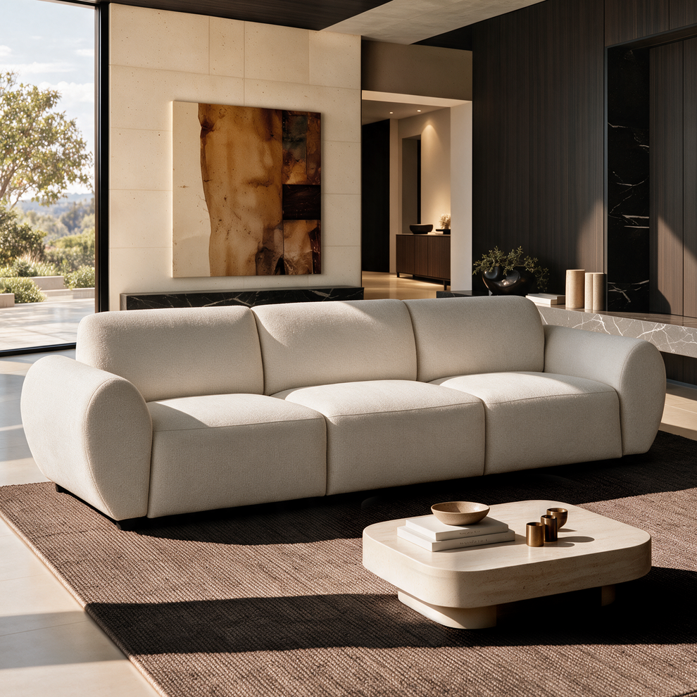 Cream-colored three-seat sofa with rounded arms in a sunlit living room, with a low coffee table, brown rug, abstract wall art and a large window.