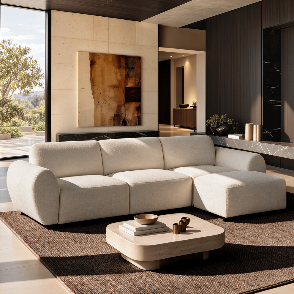 Cream sectional sofa with rounded arms and a right-side chaise in a sunlit living room, with a low coffee table, brown rug, abstract wall art and large windows.