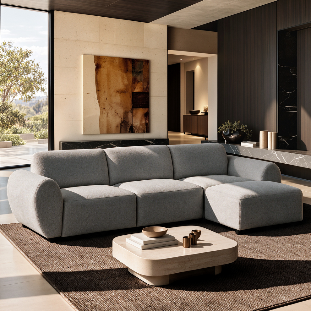 Light gray sectional sofa with rounded arms and a right-side chaise in a sunlit living room with a low cream coffee table, brown rug, abstract wall art and large window.