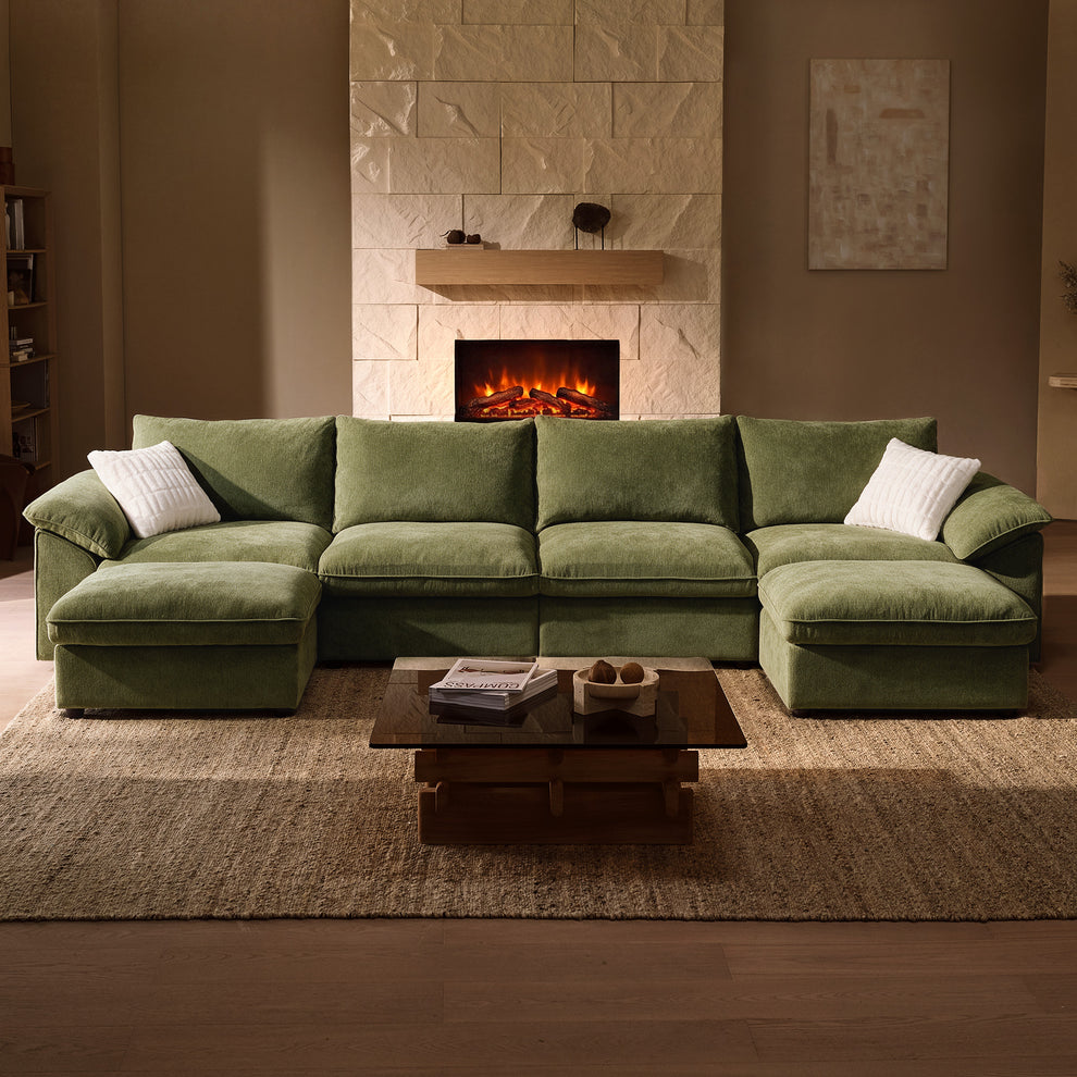 Green sofa with chaise extensions at both ends and two white pillows, facing a low coffee table on a beige rug, with a lit fireplace behind it.