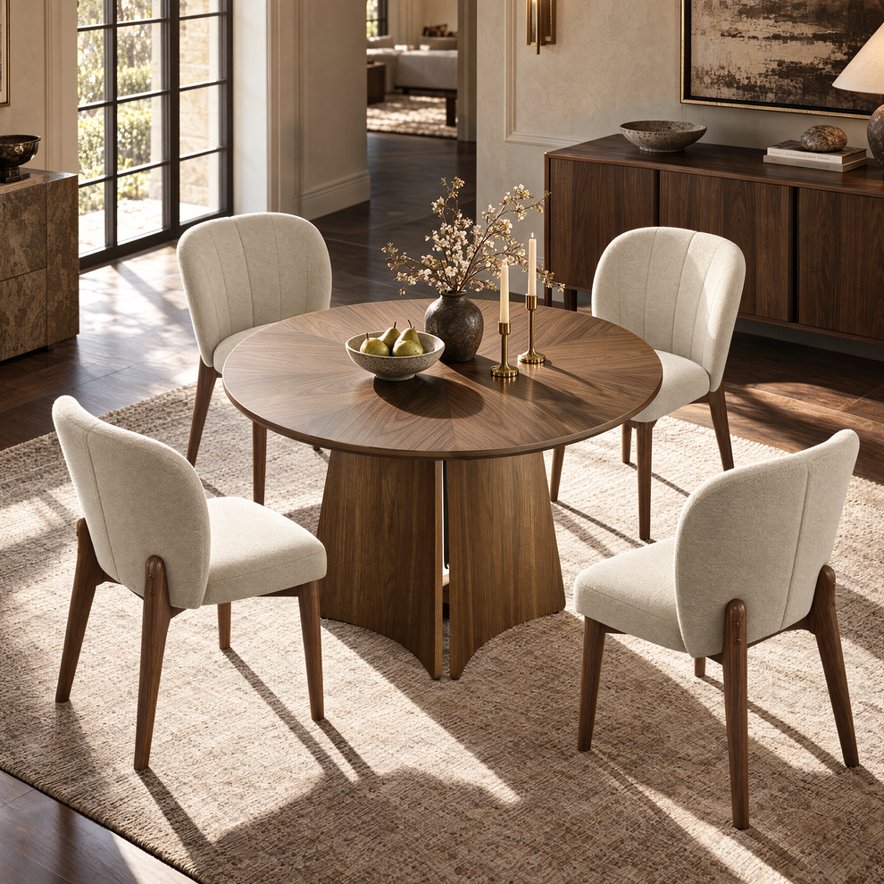 A round wooden dining table with four cream upholstered chairs is set with a bowl of pears, flowering branches, and two candles in a sunlit room.