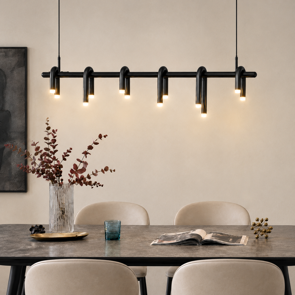 Black pendant light with six curved tubular loops and glowing tips hangs above a gray dining table with cream chairs, a vase of branches, a blue drinking glass and an open magazine.
