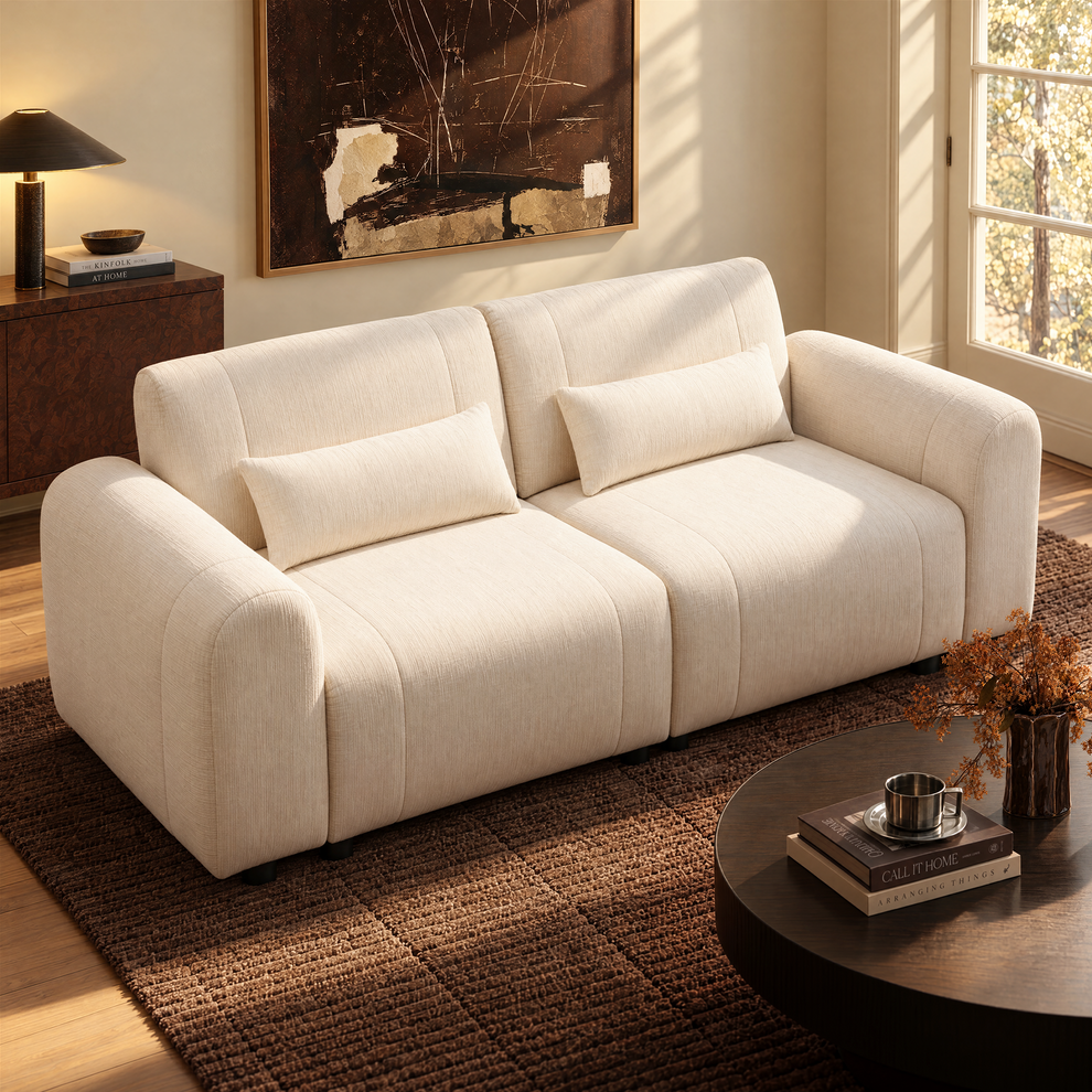 A cream-colored sofa with two matching cushions sits on a dark textured rug in a sunlit living room.