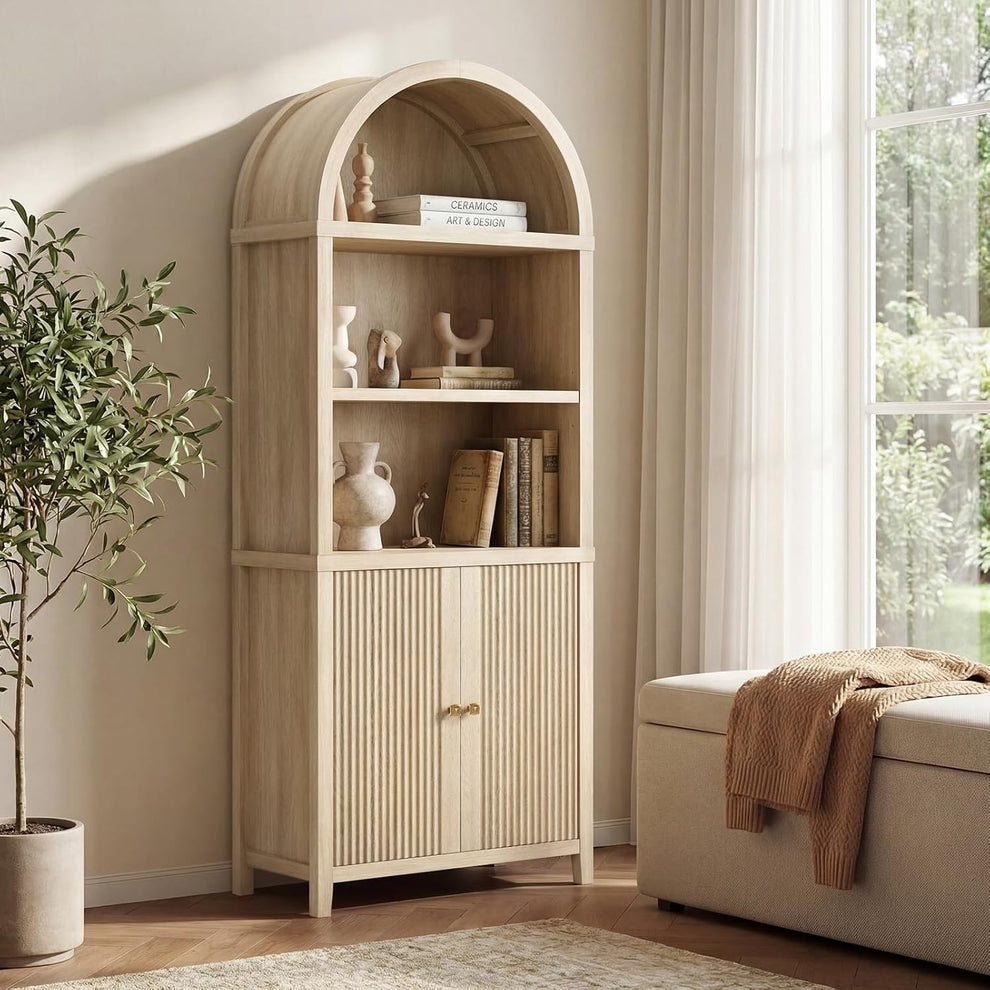 Arched pale wood-tone bookcase with open display shelves and fluted lower doors beside a window