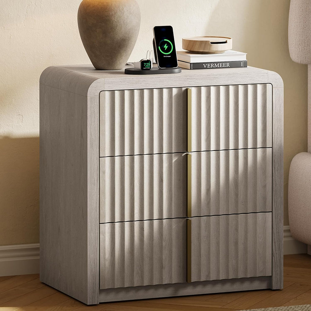 Pale wood-grain nightstand with three fluted drawers and vertical gold-tone pulls beside an upholstered bed