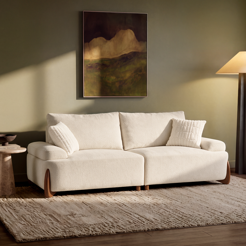 A cream sofa with two cushions sits on a textured rug beneath a framed landscape painting; a floor lamp and side table flank the scene.
