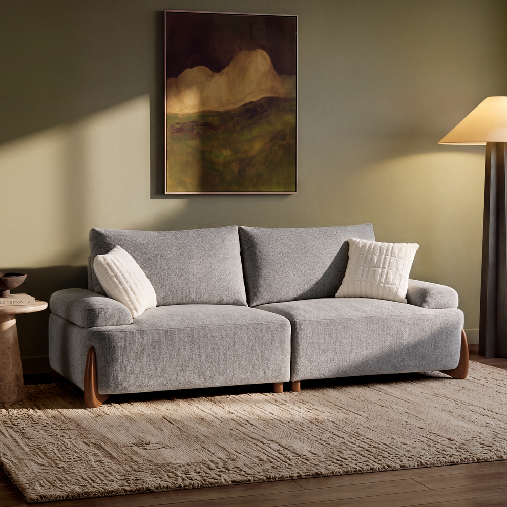 A gray sofa with two white cushions sits on a textured rug beneath a framed landscape painting, beside a floor lamp.