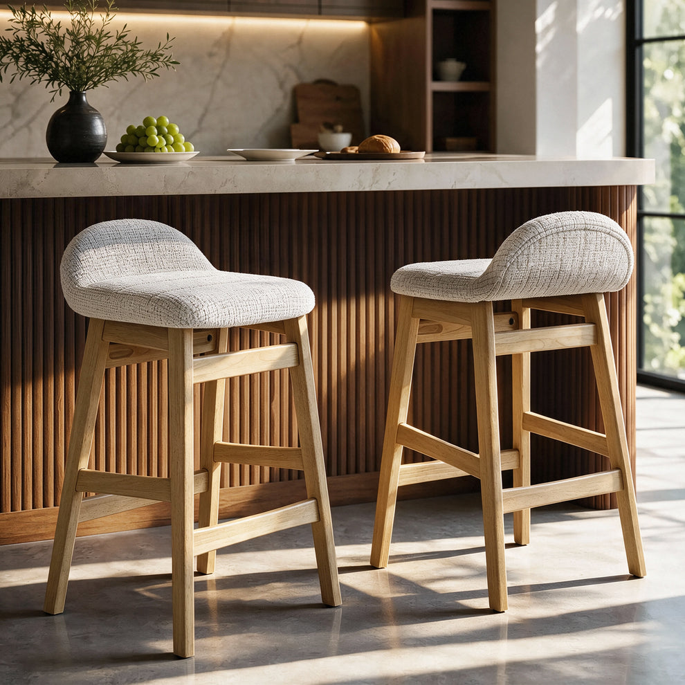 Two counter stools with cream textured seats, curved low backs and light brown legs stand beside a kitchen island with a pale countertop and dark vertical slats.