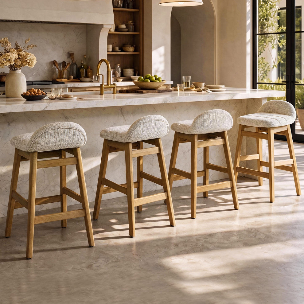 Four tall stools with cream upholstered seats, curved low backs and light brown frames line a pale veined kitchen island in a sunlit room.