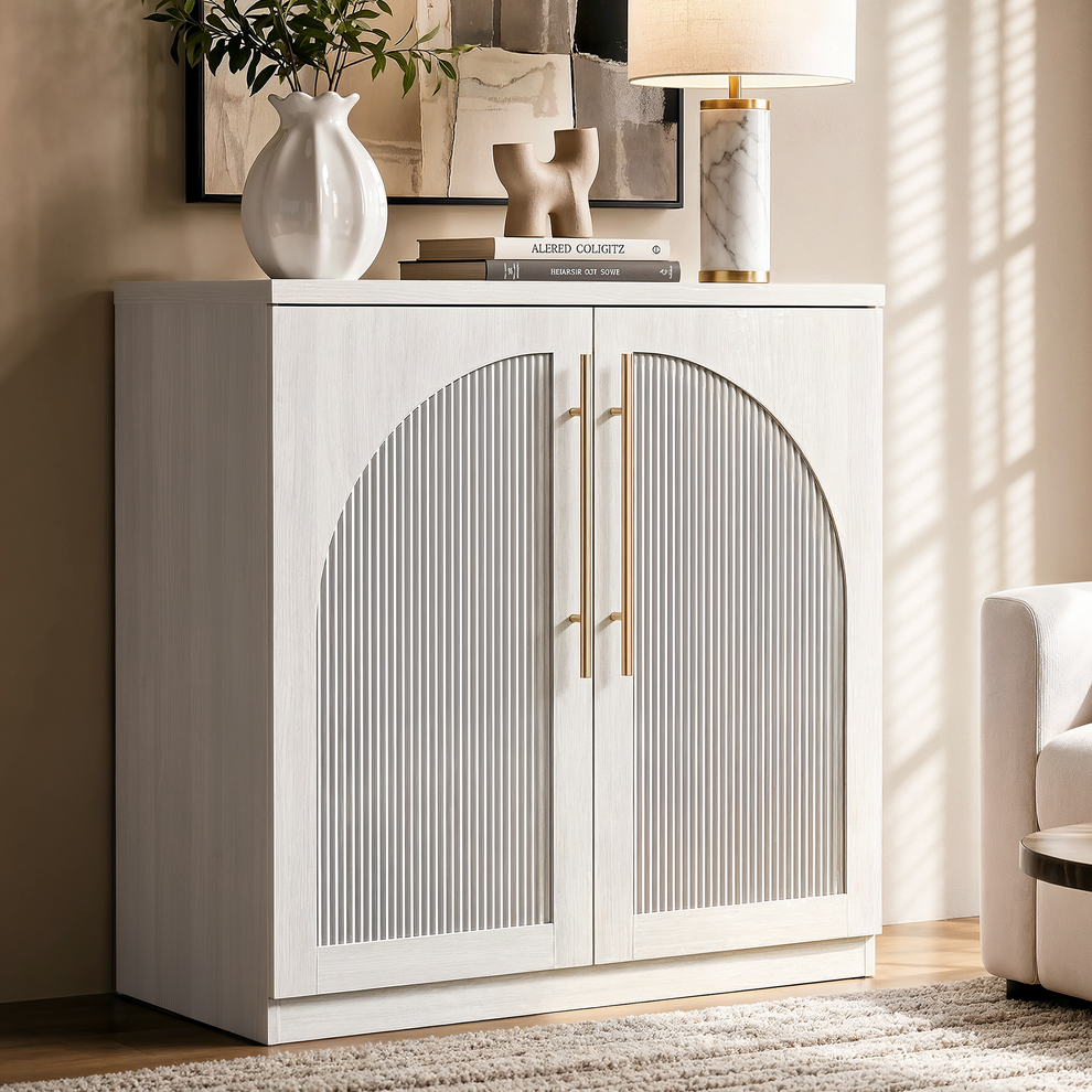 White accent cabinet with arched fluted double doors and long gold-tone handles beside a cream chair