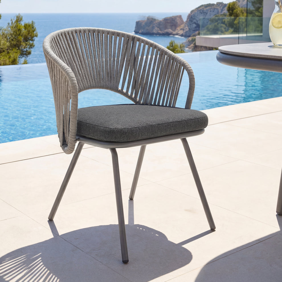 Gray cord-wrapped outdoor armchair with dark seat cushion and slender legs, Chair stands beside a pool overlooking rocky coastline