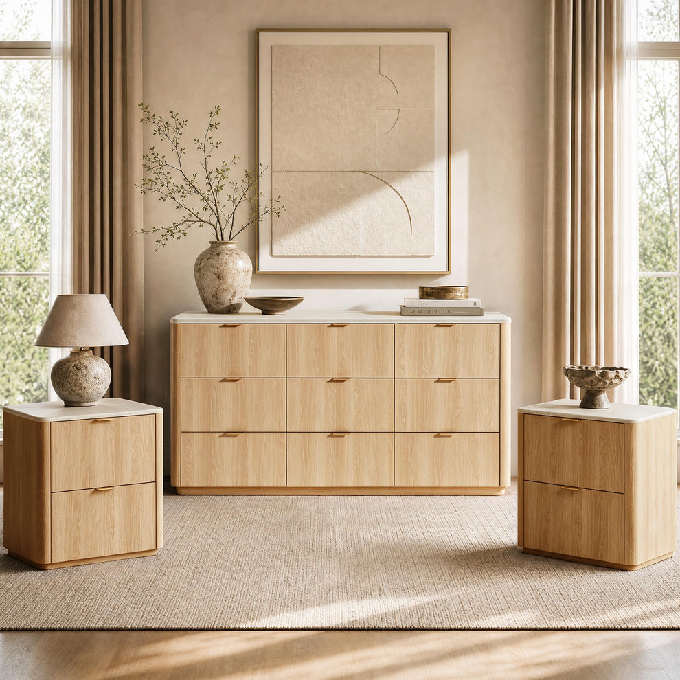 Light wood-tone nine-drawer dresser and two matching two-drawer cabinets with pale tops and gold-colored handles, arranged beneath abstract wall art with a lamp, leafy vase and bowls.