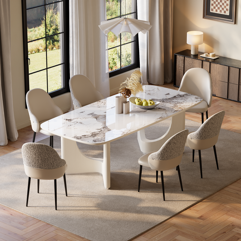 Dining room with a white veined table on fluted curved supports, six cream chairs with black legs, a bowl of green apples and vases beneath a white pendant light.
