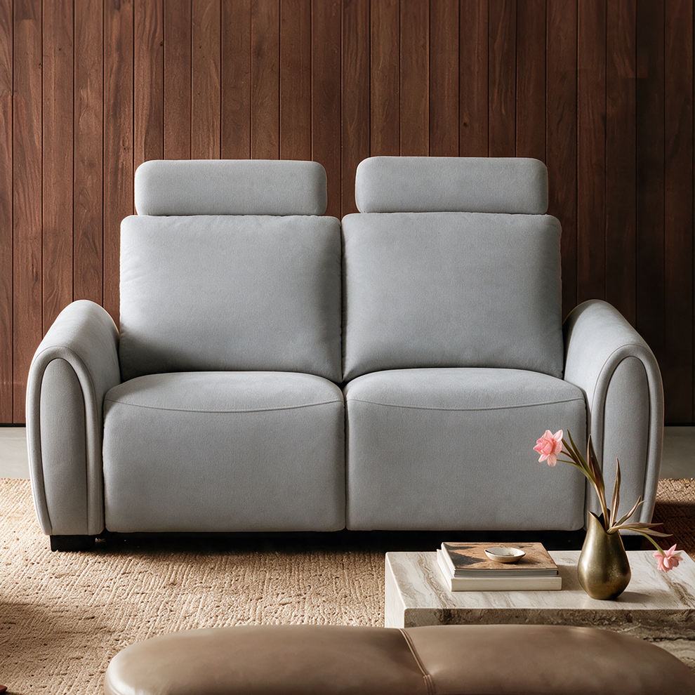 Light gray two-seat sofa with tall padded backs, rectangular headrests and curved arms, set against a brown paneled wall with a low table and pink flowers in front.
