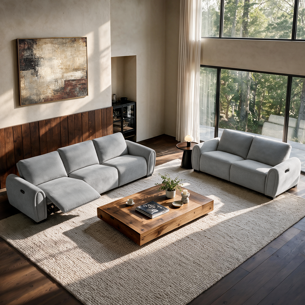 Two light gray sofas, one with an extended footrest, surround a low rectangular coffee table on a cream rug in a sunlit room with tall windows and abstract wall art.