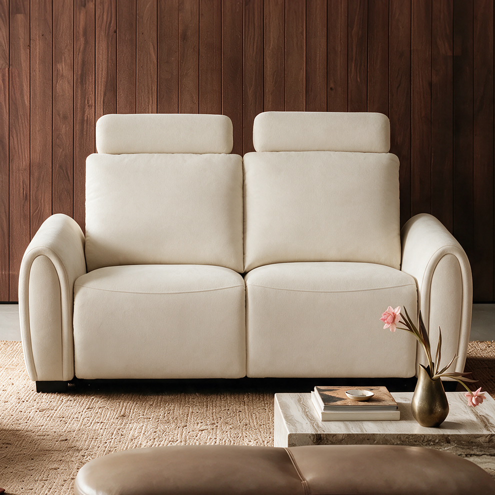 Cream two-seat sofa with tall padded backs, headrests and curved arms against a brown paneled wall, with a rug and a low table holding books and pink flowers.