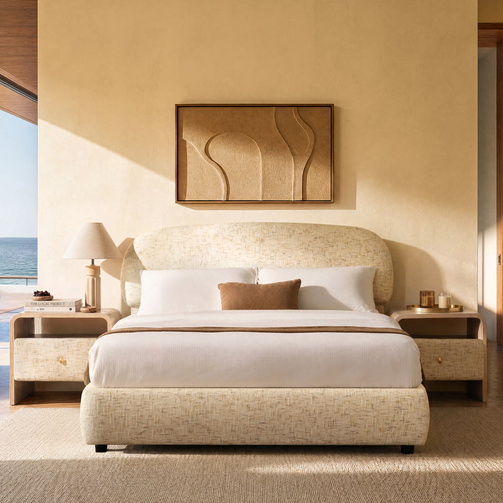 A cream upholstered bed with white bedding and a brown accent pillow stands between matching nightstands in a sunlit bedroom overlooking the sea.