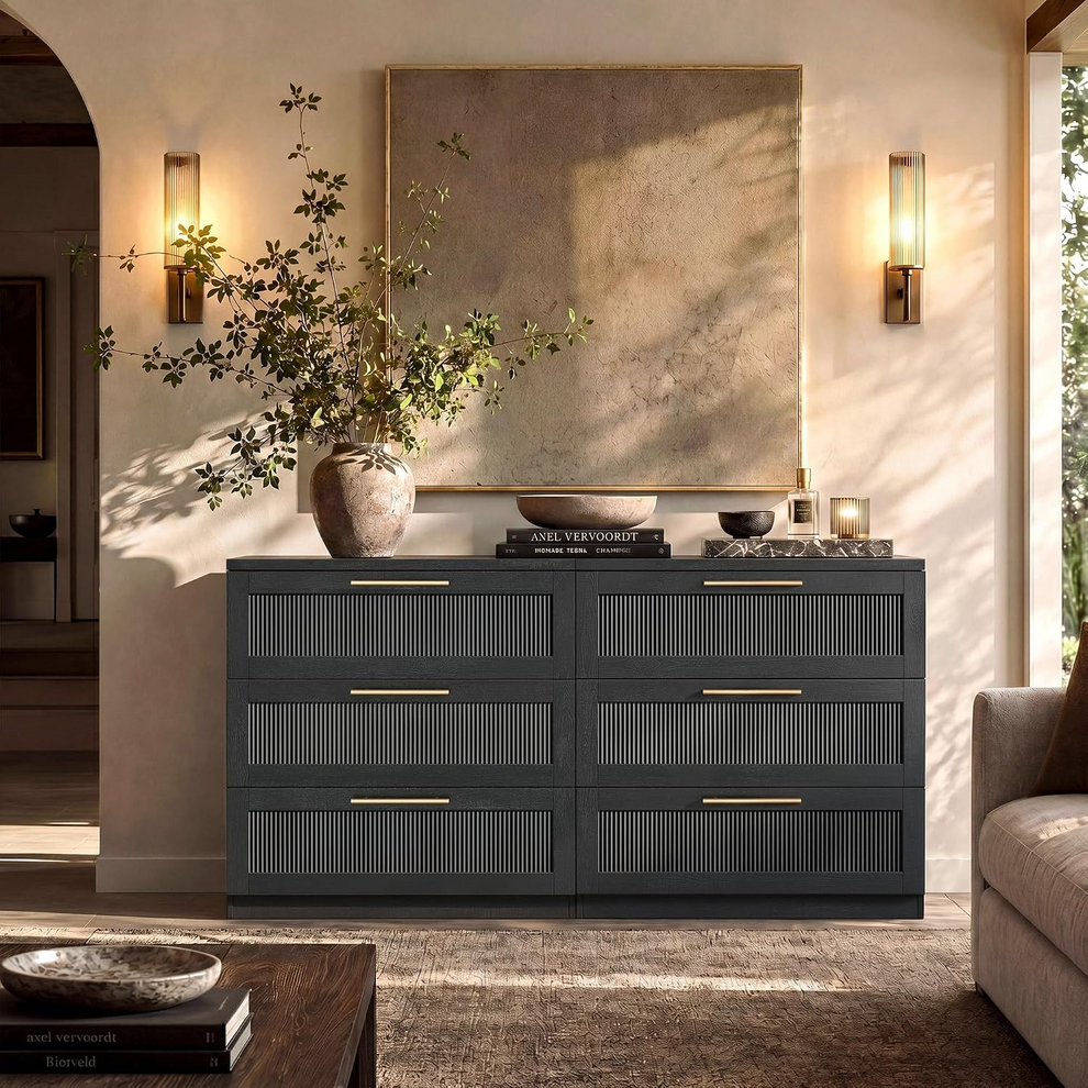 A dark six-drawer cabinet with vertical slatted fronts and brass handles stands beneath framed artwork, flanked by lit wall sconces in a sunlit living room.
