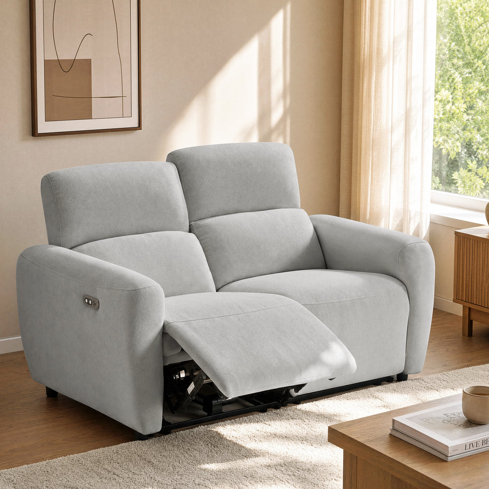Gray reclining loveseat with one footrest extended and side controls, shown beside a living room window