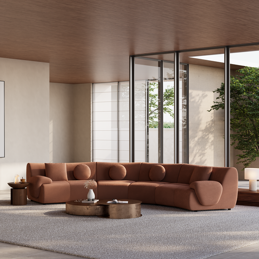 Curved rust-colored sofa with round and rectangular cushions in a bright living room with low coffee tables, a pale rug, and tall windows overlooking trees.