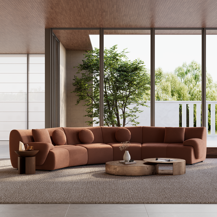 A rust-colored curved sectional sofa with round and square cushions sits behind two low coffee tables on a textured rug, beside tall windows overlooking trees.