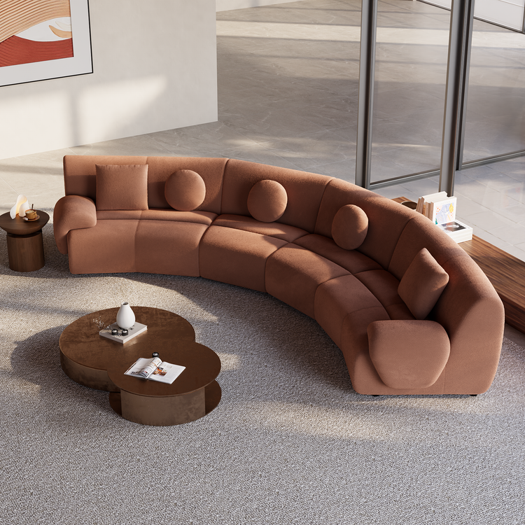 Curved rust-colored sofa with rounded arms, three round cushions and two square cushions on a light rug, beside overlapping round coffee tables in a sunlit room.