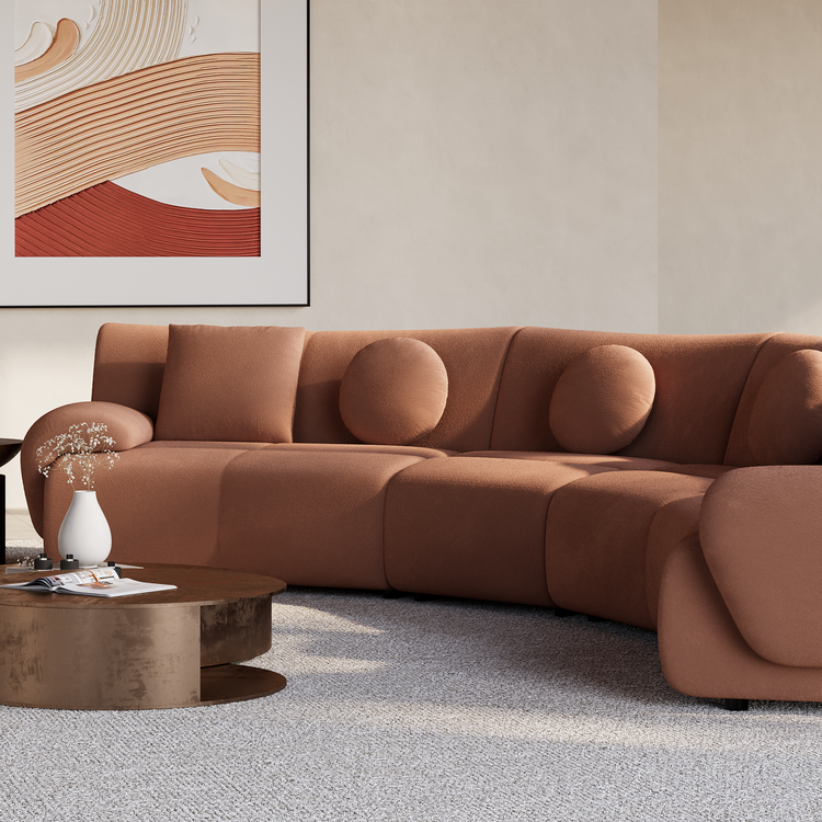 A curved rust-colored sectional sofa with round and square cushions sits on a light rug beside a low coffee table, beneath framed abstract artwork.
