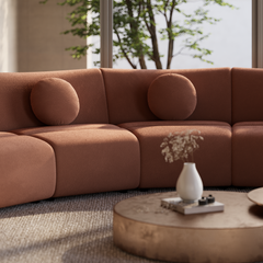 Terracotta-colored sofa with rounded seats and two circular cushions beside a low oval table holding a white vase, with leafy trees visible through windows behind.
