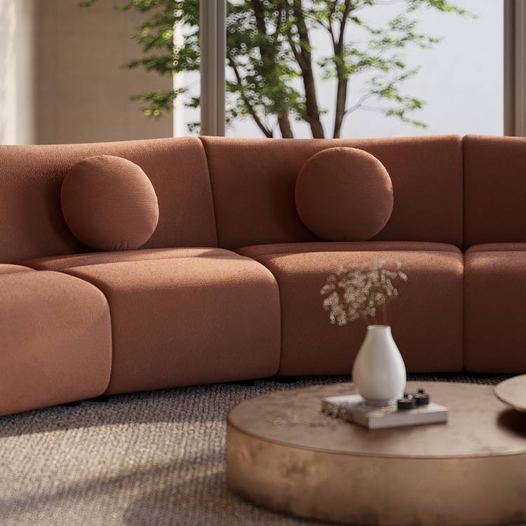 Terracotta-colored sofa with rounded seats and two circular cushions beside a low oval table holding a white vase, with leafy trees visible through windows behind.