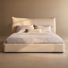 Beige upholstered bed with a tall, center-seamed headboard, pale bedding, four large pillows and a small textured rectangular cushion against a plain tan wall.
