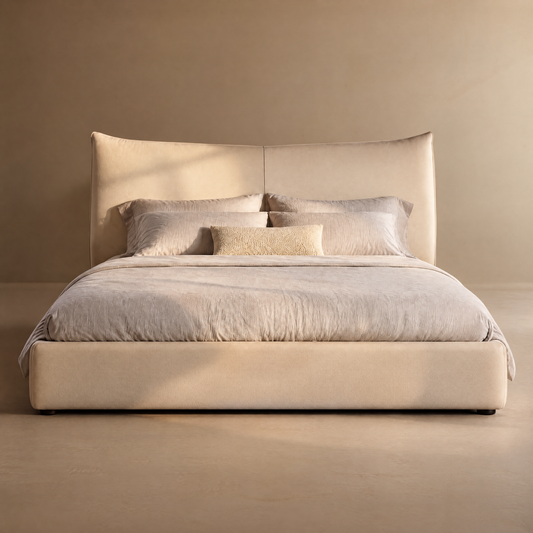 Beige upholstered bed with a tall, center-seamed headboard, pale bedding, four large pillows and a small textured rectangular cushion against a plain tan wall.