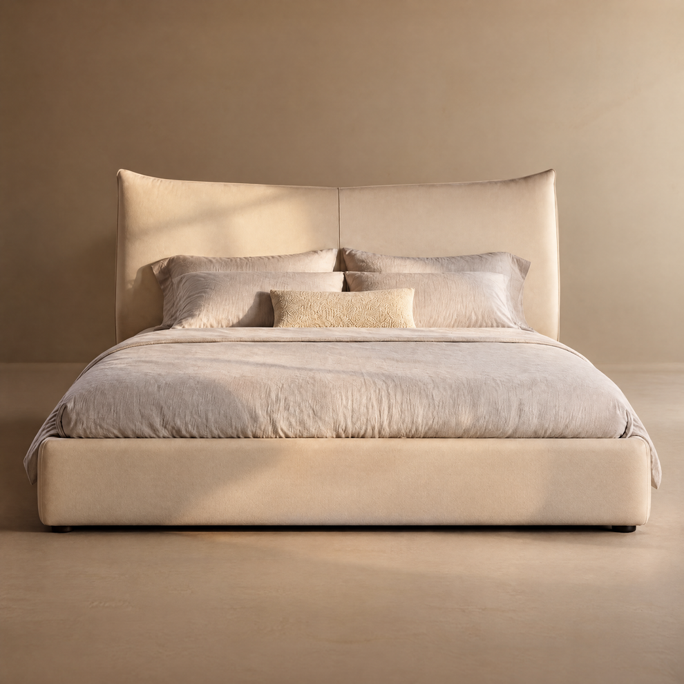 Beige upholstered bed with a tall, center-seamed headboard, pale bedding, four large pillows and a small textured rectangular cushion against a plain tan wall.