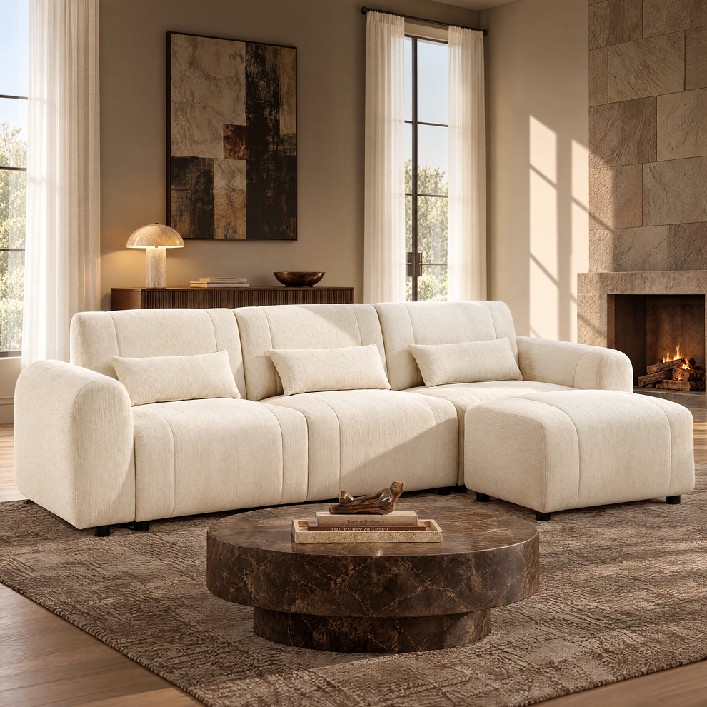 Cream sofa with three matching pillows and a rectangular ottoman in a sunlit living room with a round brown coffee table, abstract wall art and a lit fireplace.