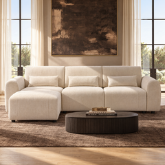 Cream sofa with rounded arms, three rectangular pillows and a chaise extending forward on the left, behind a dark oval coffee table beneath a large abstract painting.