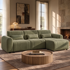An olive-green sectional sofa with three cushions and a chaise sits in a sunlit living room, with a round wooden coffee table and a stone fireplace.