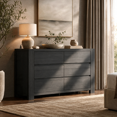 Dark gray six-drawer dresser topped with a lamp, a shallow bowl of leafy branches, books and a small vase, beneath framed artwork beside a curtained window.