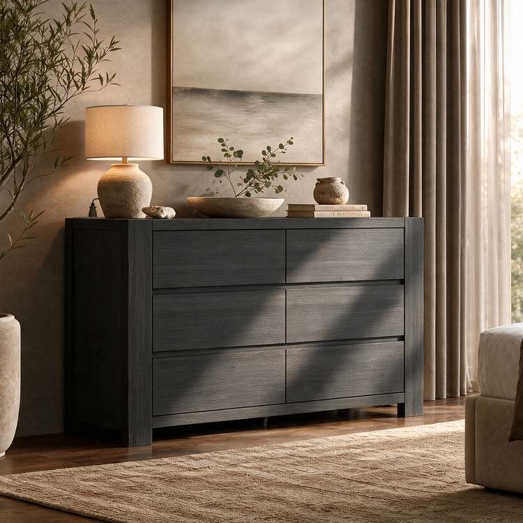 Dark gray six-drawer dresser topped with a lamp, a shallow bowl of leafy branches, books and a small vase, beneath framed artwork beside a curtained window.