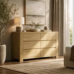Light wood-grain six-drawer chest topped with a lamp, leafy arrangement and stacked books beneath framed artwork, beside a potted tree and curtained window.