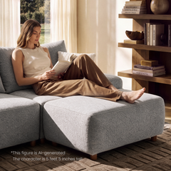 A woman reads a book while reclining barefoot on a light gray sectional sofa beside a curtained window and shelves holding books and decorative objects.