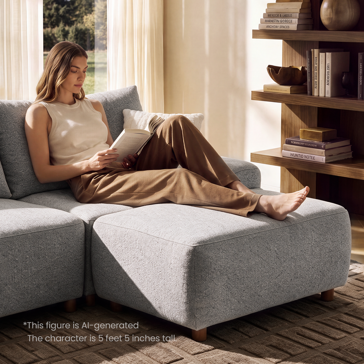 A woman reads a book while reclining barefoot on a light gray sectional sofa beside a curtained window and shelves holding books and decorative objects.