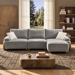 A gray sectional sofa with a chaise and two white pillows sits in a living room before a curtained window, with a wooden coffee table and bookcase nearby.