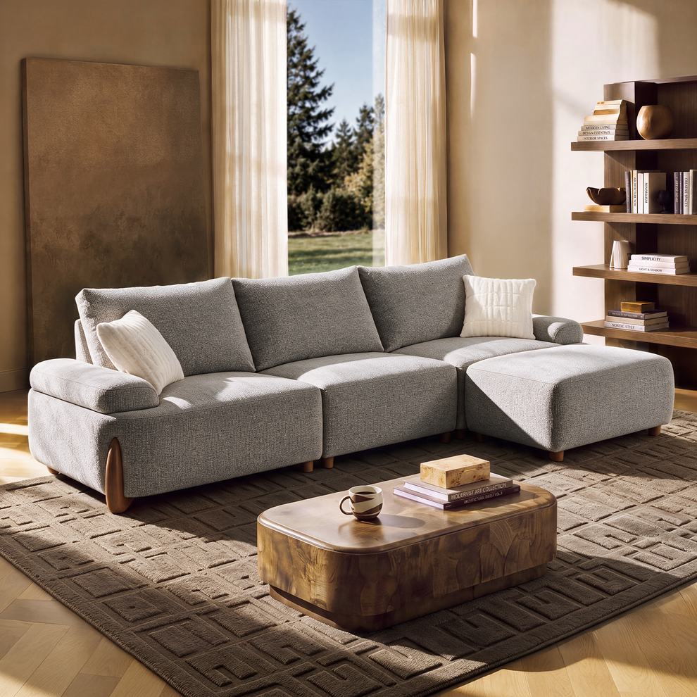A gray sectional sofa with two white cushions sits on a dark patterned rug beside a wooden coffee table in a sunlit living room.