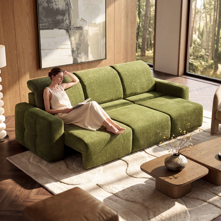 A woman holding an open book reclines on a green sofa with three extended footrests in a sunlit room with abstract wall art and large windows overlooking trees.