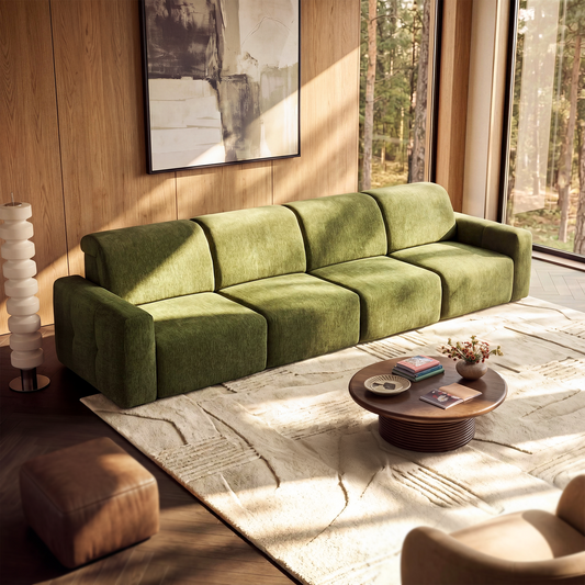 Green four-seat sofa with broad arms in a sunlit room, with a cream patterned rug, round coffee table, abstract wall art and tall windows overlooking trees.
