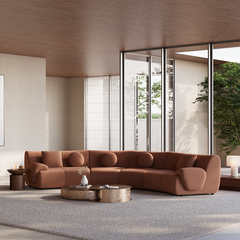 Curved rust-brown sofa with round and square cushions faces two low coffee tables on a pale rug in a bright room with floor-to-ceiling windows.